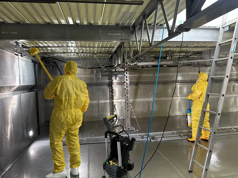 Zwei Mitarbeiter in gelben Schutzanzügen reinigen Wände und Mechanik eines leeren Edelstahlbeckens